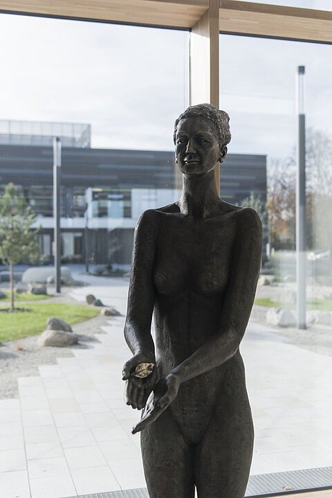 Eine bronzene Skulptur einer nackten Frau steht im Vordergrund. Sie hat die Arme leicht ausgebreitet, die Hände sind offen. Im Hintergrund sind Fenster und ein grüner Außenbereich mit Pflanzen und Steinen sichtbar.
