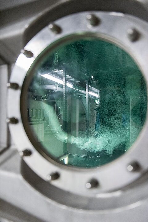 Ein runder Metallrahmen mit einer grünen Glasscheibe bietet einen Blick auf ein Innenleben mit Rohren und Wasser, das aufsteigt und blubbert. Die Umgebung wirkt industriell und gut beleuchtet.