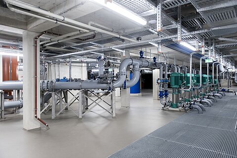 Eine industrielle Rohrleitungsanlage in einem großen Gebäude. Sichtbare Rohre verlaufen entlang der Wände und Decke, unterbrochen von Pumpen und Ventilen. Der Boden ist aus grauem Material, und das gesamte Umfeld wirkt modern und gut beleuchtet.