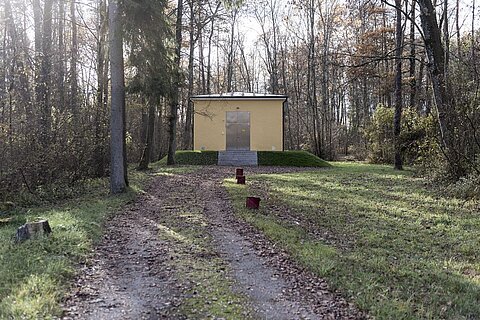 Ein gepflasterter Weg führt durch einen Wald zu einem gelben Gebäude, das auf einer kleinen Plattform steht. Umgeben von Bäumen und Sträuchern sind auf dem Weg einige rote Behälter sichtbar. Die Szenerie wirkt ruhig und abgelegen.