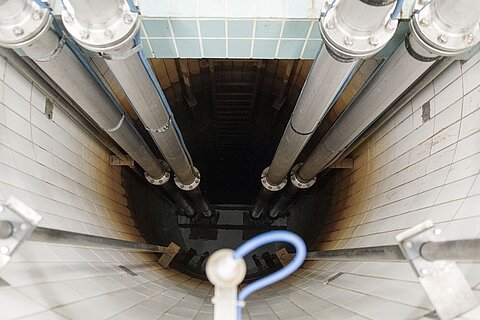 Blick auf einen tiefen Schacht mit mehreren großen Rohren, die vertikal nach unten führen. Der Schacht ist gefliest und am Boden ist dunkles Wasser zu sehen. Ein blauer Schlauch ist sichtbar, der sich um ein Metallteil windet.
