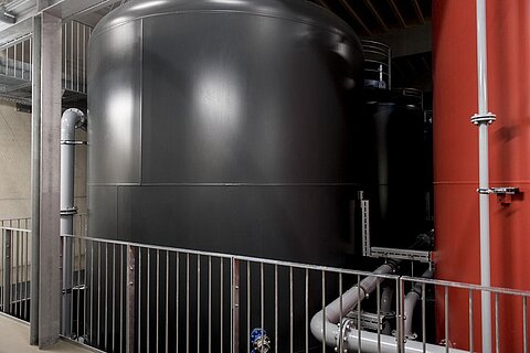 Ein großer, schwarzer Tank steht neben einer rot gestrichenen Wand, umgeben von Metallgeländern und Rohren. Der Raum hat eine industrielle Atmosphäre mit sichtbaren Rohrleitungen, die den Tank verbinden.