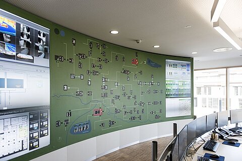 Ein Kontrollraum mit einer großen, interaktiven Wandkarte zeigt verschiedene Verbindungen und Datenpunkte. Screens zeigen zusätzliche Informationen und Grafiken. Der Raum ist modern und hell mit mehreren Arbeitsplätzen für Analysten.