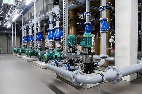 Eine Reihe von großen Rohren und Pumpen in einem technischen Raum. Die Rohrleitungen sind in verschiedenen Farben, mit grünen und blauen Ventilen. Die Wände sind aus Beton, und die Beleuchtung ist hell, was den industriellen Charakter des Raums unterstreicht.
