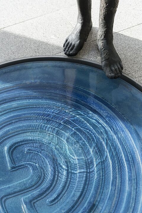 Eine Skulptur mit einer menschlichen Figur steht neben einem runden Wasserbecken. Das Wasser zeigt konzentrische Linien, die wie ein Labyrinth wirken. Die Füße der Figur sind barfuß und stehen nahe am Rand des Beckens.