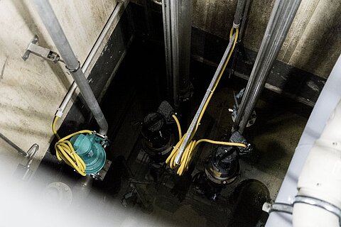 Blick in einen sumpfigen Raum mit mehreren Pumpen, die in Wasser stehen. An den Pumpen sind gelbe Kabel befestigt, die an vertikale Rohre angeschlossen sind. Die Wände des Raumes sind steinig und es gibt wenig Licht.