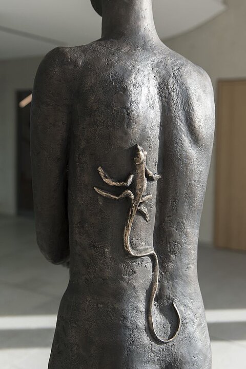 Eine Skulptur eines menschlichen Rückens, auf dem eine kleine Echse platziert ist. Die Figur ist dunkel und hat eine glatte Oberfläche, während die Echse hell und detailliert gestaltet ist. Der Hintergrund zeigt einen modernen Raum.