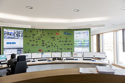 Ein Kontrollraum mit mehreren Bildschirmen, die auf eine digitale Übersichtskarte zeigen. Ein Mitarbeiter sitzt links und überwacht die Daten. Der Raum hat große Fenster, die Tageslicht hereinlassen, und die Wände sind modern gestaltet.