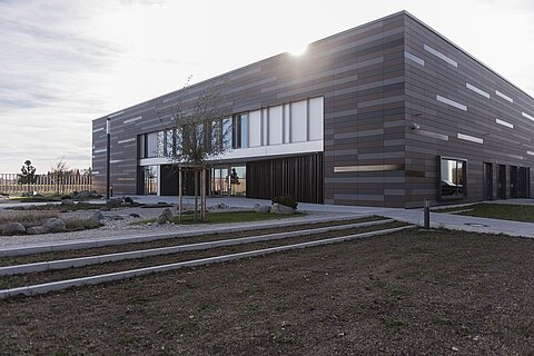 Moderne Gebäude mit einer Fassade aus braunen und grauen Paneelen. Es gibt große Fenster und eine ansprechende Landschaftsgestaltung mit Steinen und Pflanzen im Vordergrund. Der Himmel ist klar und die Sonne scheint auf das Gebäude.
