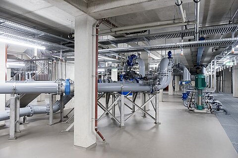 Ein insgesamt technisches Bild eines modernen Rohrsystems in einer Industrieanlage. Graue Rohre verlaufen entlang der Wände, mit Ventilen und Anschlussstücken. Im Hintergrund sind verschiedene Maschinen und eine grüne Komponente sichtbar. Die Umgebung ist sauber und gut beleuchtet.