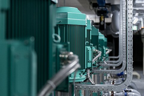 Mehrere grüne Elektromotoren sind in einer industriellen Umgebung aufgereiht, verbunden durch Rohre und Leitungen. Die Geräte befinden sich in einem gut beleuchteten Raum mit sichtbaren technischen Installationen.
