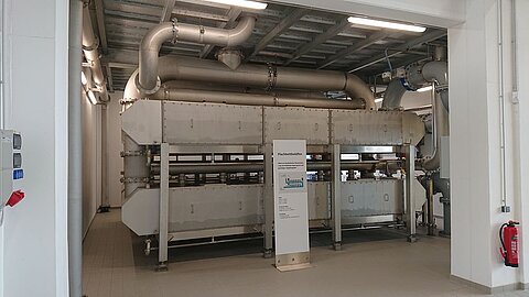 Eine große Maschinenanlage steht in einem hellen Raum. Sie ist mit mehreren Rohren ausgestattet, die von der Decke herabführen. An der Seite befindet sich ein Informationsschild. Der Boden ist hell und sauber.