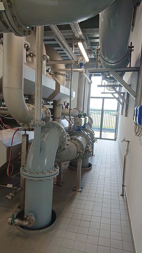Ein Industriebereich mit großen Rohren und Ventilen, die an einer Wand entlang verlaufen. Im Hintergrund ist ein Fenster mit natürlichem Licht sowie ein Treppenaufgang zu sehen. Die Böden sind gefliest und die Wände sind hell.