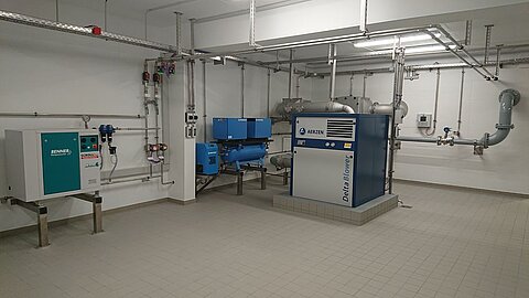 Ein technischer Raum mit zwei Maschinen. Links steht ein kleinerer Gerät mit einer grünen und weißen Oberfläche, rechts eine größere Maschine mit blauer und grauer Haupteinheit. Rohre und Ventile verlaufen an den Wänden und Decke. Der Boden ist gefliest und der Raum wirkt gut beleuchtet.