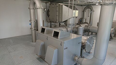 Nahaufnahme eines technischen Raums mit großen Metallrohren und einem zentralen, rechteckigen Gerät. Der Boden ist mit rutschfestem Material ausgelegt. Die Wände sind schlicht und hell. Die Einrichtung scheint für industrielle Prozesse konzipiert zu sein.