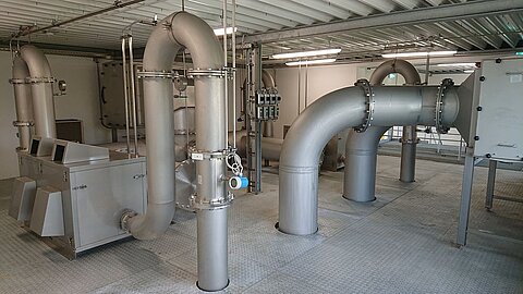 In einem industriellen Raum sind große silberne Rohre installiert, die durch den Raum verlaufen. An einer Wand befindet sich eine Maschinenanlage. Der Boden ist mit Metallplatten ausgelegt. Die Beleuchtung sorgt für eine helle Umgebung.