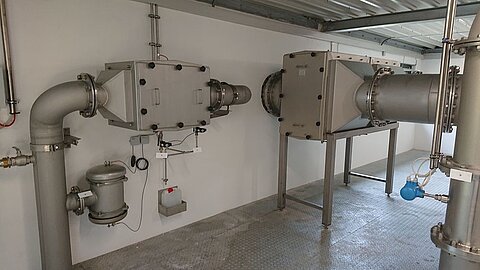Das Bild zeigt eine Fabrikverrohrung mit zwei großen metallenen Behältern, die durch Rohre verbunden sind. Im Vordergrund sind verschiedene Ventile und Instrumente sichtbar. Die gesamte Installation befindet sich in einem Raum mit einem grauen Boden und einer gefliesten Decke.