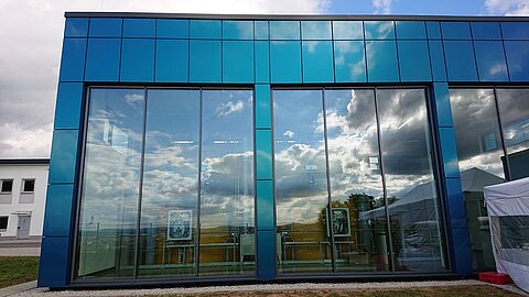 Die Vorderseite eines modernen Gebäudes mit großen Glasfenstern, die den Himmel und Wolken spiegeln. Die Fassade ist in einem blauen Farbton gestaltet, und der Untergrund zeigt einen kleinen Bereich mit Rasen und Kies.