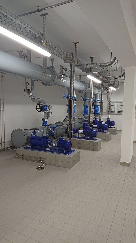 In einem Raum sind mehrere blaue Pumpen und Rohre sichtbar. Die Pumpen sind auf Plattformen montiert und mit Steuer- und Regelventilen ausgestattet. Eine helle Beleuchtung sorgt für gute Sicht. Der Boden ist gefliest.