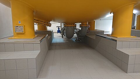 Ein Innenraum mit einem großen, gelben Behälter, der auf massiven Stützen steht. Darunter verlaufen Rohre und der Boden ist gefliest. An den Wänden sind technische Einbauten sichtbar. Das Licht ist hell und der Raum wirkt sauber und funktional.