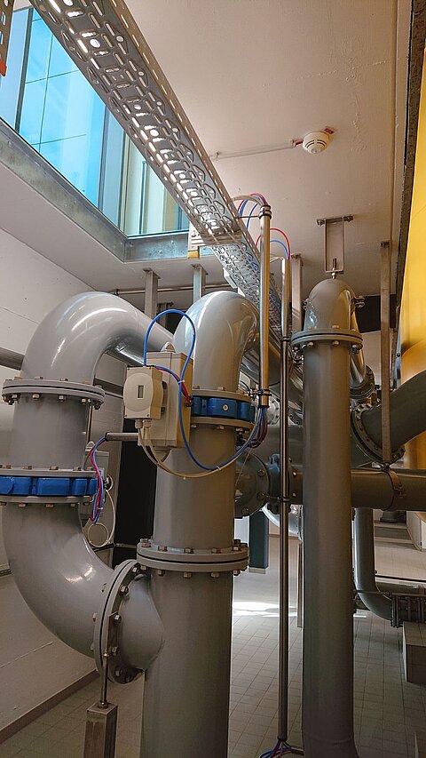 Das Bild zeigt eine industrielle Umgebung mit mehreren großen grauen Rohren, die miteinander verbunden sind. Im Hintergrund sind Fenster vorhanden, durch die natürliches Licht fällt. Die Rohre sind mit verschiedenen elektrischen Leitungen und Anschlüssen ausgestattet.