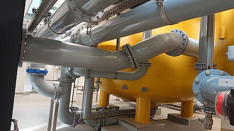 Das Bild zeigt eine industrielle Anlage mit großen, gelben Behältern und zahlreichen Rohren in verschiedenen Größen, die miteinander verbunden sind. Die Rohre sind in Grautönen gehalten und verlaufen an der Decke und entlang des Bodens der Anlage.
