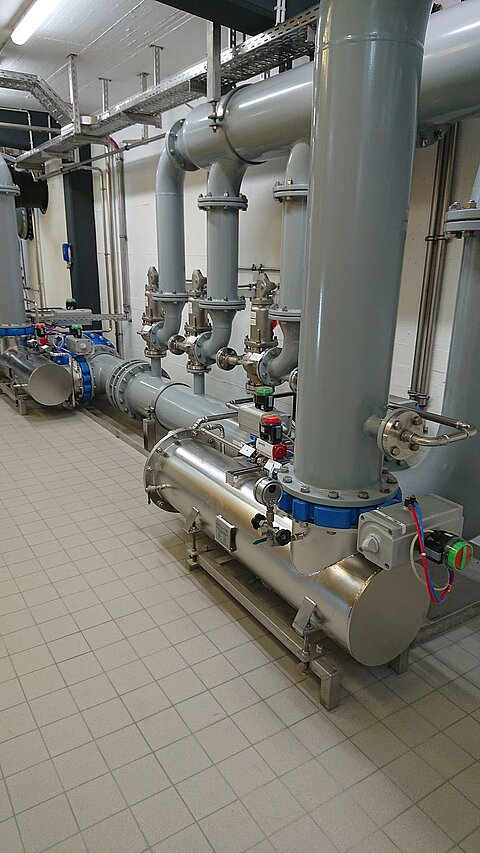 In einem Raum sind mehrere große Metallrohre und Ventile zu sehen, die zur industriellen Infrastruktur gehören. Die Rohre sind grau und mit Armaturen ausgestattet. Der Boden ist gefliest und der Raum wirkt ordentlich und gut organisiert.