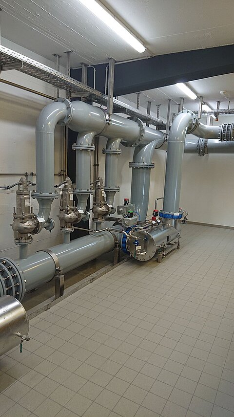 Das Bild zeigt ein Rohrleitungsystem in einer industriellen Umgebung. Mehrere große graue Rohre sind installiert, begleitet von Ventilen und Anschlüssen auf Fliesenboden. Die Beleuchtung ist hell und gleichmäßig.