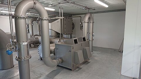 Ein industrieweißer Raum mit großen metallischen Rohren und einem Ausrüstungsgehäuse. Die Rohre sind glatt und verankert, die Wände sind neutral gehalten. Der Boden ist strukturiert und reflektiert das Licht, was auf eine funktionale Anlage hinweist.