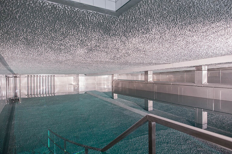 Eine moderne Innenansicht eines Raums mit glatten, reflektierenden Oberflächen und einem Pool. Die Beleuchtung schafft eine kühle Atmosphäre, während das Wasser des Pools den Raum teilweise spiegelt. Eine Treppe führt ins Wasser, und im Hintergrund ist eine metallische Struktur sichtbar.