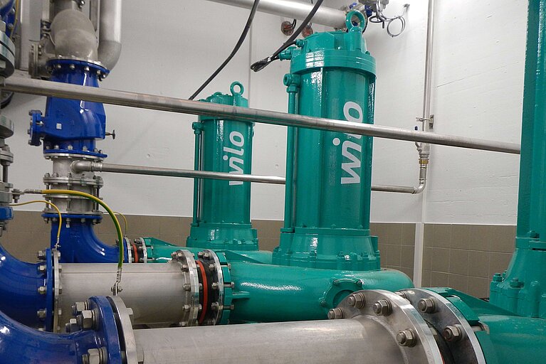Das Bild zeigt eine industrielle Pumpenanlage mit mehreren grünen Pumpen und blauen Rohren. Die Rohre sind aus Metall und verfügen über Flansche, die mit Schrauben befestigt sind. Die Wand im Hintergrund ist hell und teilweise gefliest.