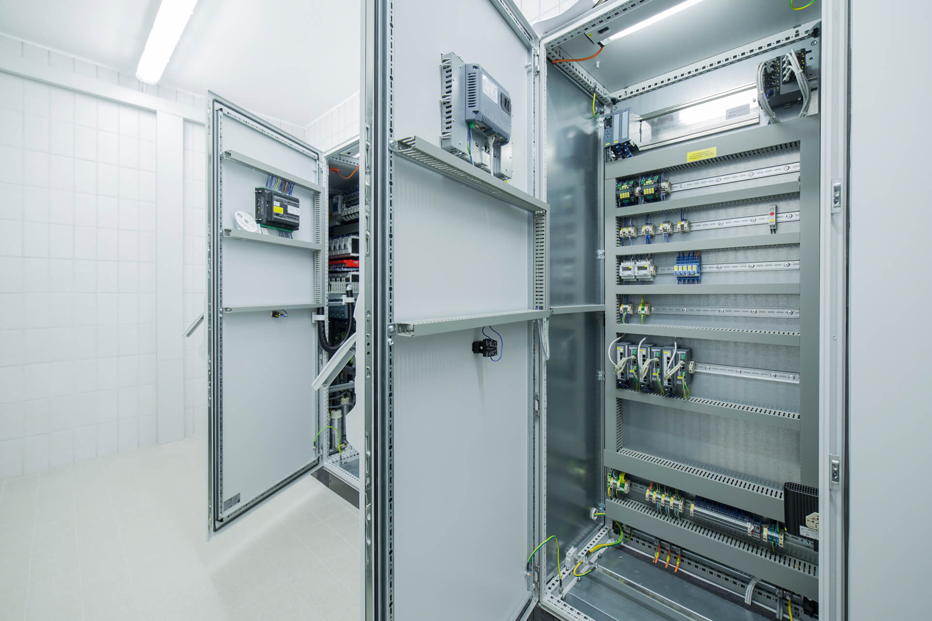 Das Bild zeigt einen Blick in einen technischen Raum mit offenen Schaltschranktüren, die elektrische Komponenten und Verkabelungen enthalten. Die Wände sind weiß gefliest und die Beleuchtung ist hell.