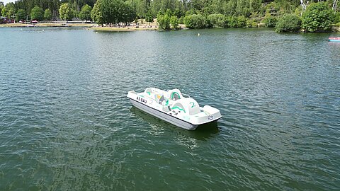 Ein Tretboot schwimmt auf einem ruhigen Gewässer, umgeben von Bäumen und einer aufgeräumten Parklandschaft im Hintergrund. Es gibt keine Menschen auf dem Boot, und die Wasseroberfläche hat sanfte Wellen.
