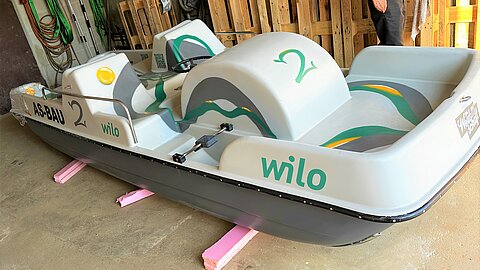 Ein graues Tretboot mit grünenund gelben Akzenten steht auf pinkfarbenen Stützen. Es trägt die Aufschrift "ASBAU 2" und "wilo". Im Hintergrund sind Holzpaletten zu sehen.