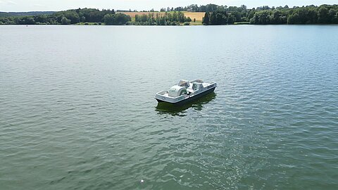 Ein kleines Boot fährt auf einem ruhigen Gewässer. Im Hintergrund sind Bäume und Hügel zu sehen, die die Landschaft umgeben. Der Himmel ist blau und das Wasser spiegelt die Umgebung wider.