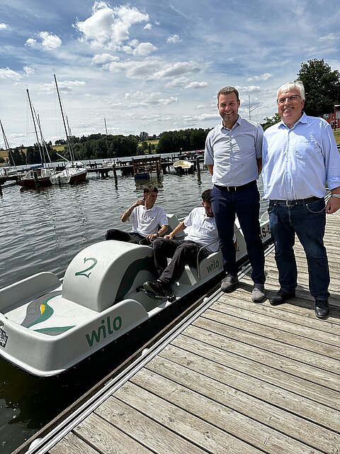 Drei Männer stehen am Ufer eines Gewässers neben einem Fahrzeug, das wie ein Tretboot aussieht. Im Hintergrund sind Segelboote und ein Holzsteg zu sehen. Der Himmel ist partly cloudy.