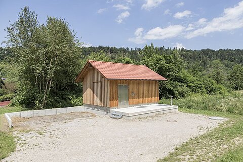 Ein kleines Holzhaus mit rotem Satteldach steht auf einem gepflasterten Platz. Umgeben von Bäumen und Grünflächen, befindet es sich in einer ländlichen Umgebung. Der Boden besteht aus Schotter, und im Hintergrund sind Hügel sichtbar.