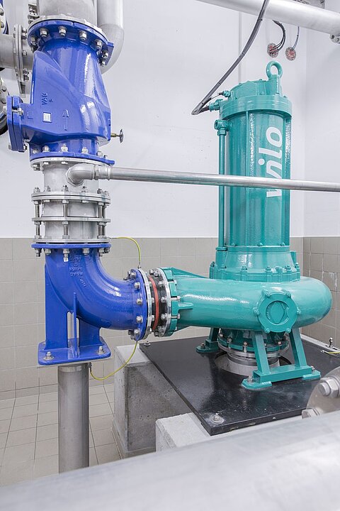 Ein blaues Rohrsystem und eine grüne Pumpe in einer industriellen Umgebung. Die Pumpe ist auf einer Plattform montiert, umgeben von Metallrohren und einem gefliesten Boden. Die gesamte Anordnung deutet auf eine Wasser- oder Abwasseranlage hin.