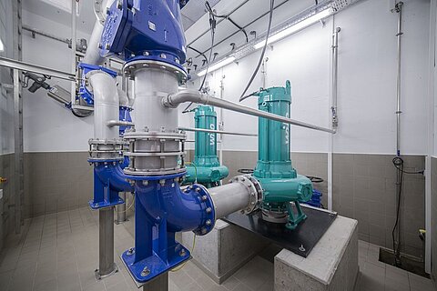 In einem mechanischen Raum sind mehrere Pumpen und Rohrleitungen zu sehen. Die Pumpen sind grün, während die Rohre blau und weiß sind. Der Boden besteht aus Fliesen, und die Wände sind grau. Die Beleuchtung ist hell und gleichmäßig.