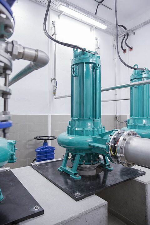 In einem technischen Raum stehen mehrere grüne Pumpen auf einer Betonplatte. Die Pumpen sind an Rohre angeschlossen, die zur Wand führen. Im Hintergrund ist ein blauer Wasserhahn sichtbar. Die Beleuchtung ist hell und die Wände sind grau gefliest.