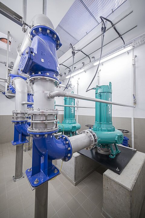 Das Bild zeigt eine industrielle Pumpenstation mit zwei grünen Pumpen, die auf einer Betontafel montiert sind. Mehrere Rohre und Ventile, hauptsächlich in blau und silber, verlaufen in verschiedenen Richtungen. Die Umgebung ist hell und modern gestaltet.