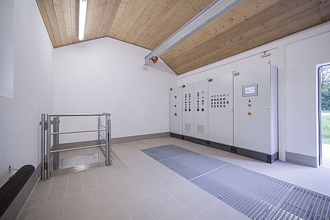 Ein Innenraum mit einer Holzdecke, der Steuerungseinheiten an einer Wand zeigt. Es gibt eine schlichte Gruppe von Schaltern und Displays. Auf dem Boden befindet sich eine Rinne und eine Treppe. Der Raum ist hell und minimalistisch gestaltet.