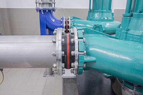 Nahtlose Verbindung von Rohrleitungen mit grünen und blauen Armaturen in einer industriellen Umgebung. Die Rohrabschnitte sind mit Flanschen und Dichtungen gesichert. Der Boden ist gefliest.