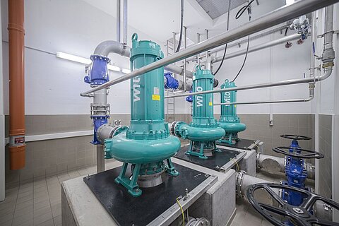 Drei grüne Pumpen stehen in einem technischen Raum, umgeben von Rohren und Ventilen. Die Pumpen sind auf schwarzen Plattformen installiert, und die Wände sind hell gefliest. Beleuchtung sorgt für eine gut sichtbare Umgebung.
