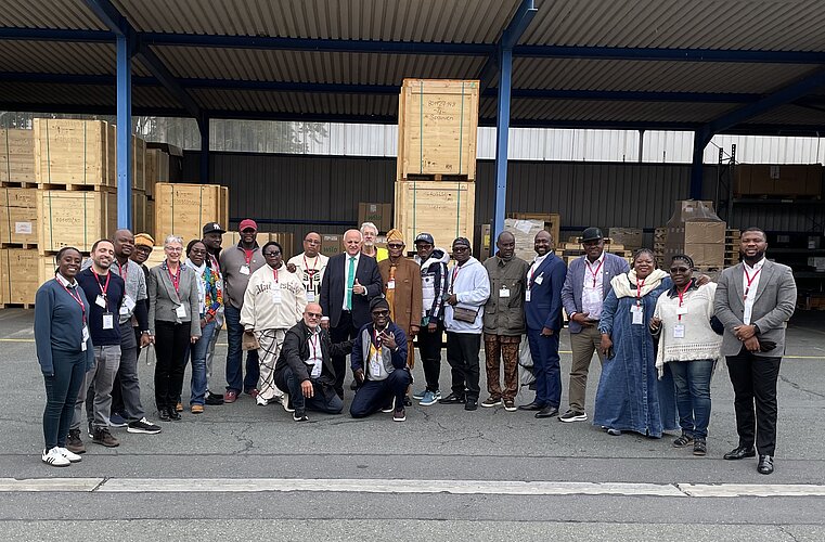 Eine Gruppe von etwa 30 Personen steht in einer Lagerhalle, umgeben von großen Holzpaletten. Die Mitglieder tragen unterschiedliche Outfits und einige haben Ausweise um den Hals. Im Hintergrund sind weitere Paletten zu sehen.