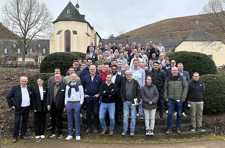 Eine große Gruppe von Menschen steht auf Treppen vor einem Gebäude und Hügeln. Die Personen sind in verschiedenen Kleiderstilen gekleidet. Die Szene wirkt freundlich und zeigt eine gemeinschaftliche Atmosphäre. Es ist bewölkt, doch die Gruppe ist gut gelaunt.