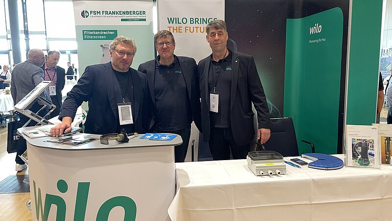 Drei Personen stehen am Stand eines Unternehmens namens Wilo auf einer Veranstaltung. Der Stand ist mit einem Banner und Produktinformationen ausgestattet. Im Hintergrund sind weitere Besucher und Stände zu sehen. Es scheint eine professionelle Umgebung zu sein.