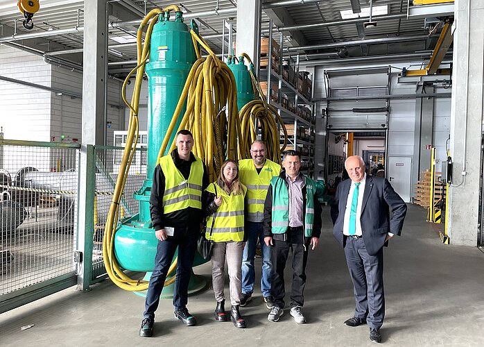 Eine Gruppe von fünf Personen steht in einer Lagerhalle mit großen grünen Maschinen im Hintergrund. Alle tragen Sicherheitswesten. Die Menschen lächeln und scheinen sich in einer beruflichen Umgebung zu befinden.