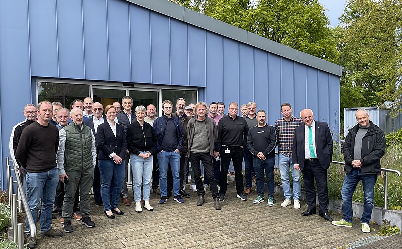 Gruppenfoto von rund 30 Personen vor einem blauen Gebäude. Die Personen stehen in mehreren Reihen, lächeln und sind unterschiedlich gekleidet. Im Hintergrund sind Bäume und eine Grünfläche sichtbar.