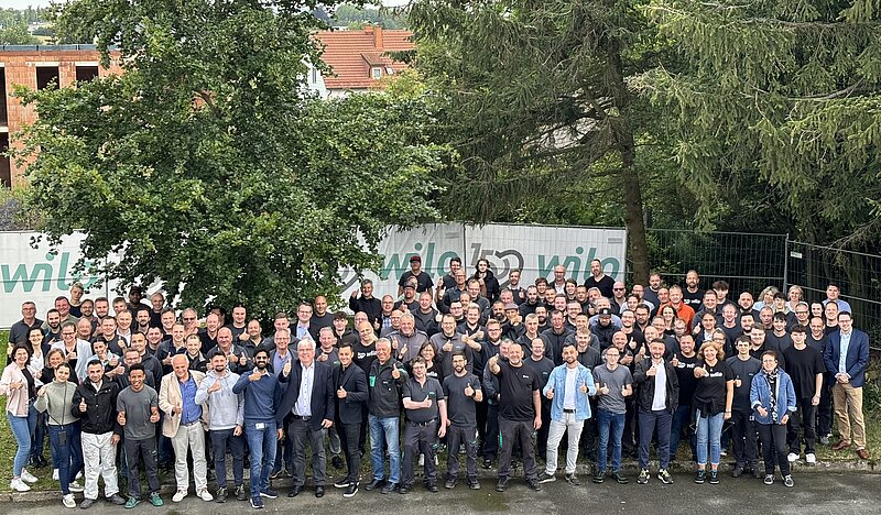 Gruppenfoto von etwa 150 Personen im Freien, versammelt vor einem Baum und einem Banner mit der Aufschrift "wild". Die Mitglieder lächeln und zeigen Daumen hoch. Die Mehrheit trägt dunkle Kleidung, einige haben helle Hemden oder Jacken an.